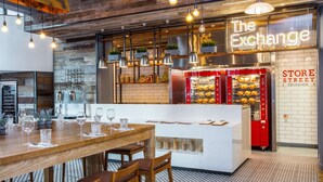 L’intérieur d’un restaurant moderne, avec une table à manger en bois, des suspensions lumineuses et un panneau néon sur lequel on peut lire « The Exchange ».