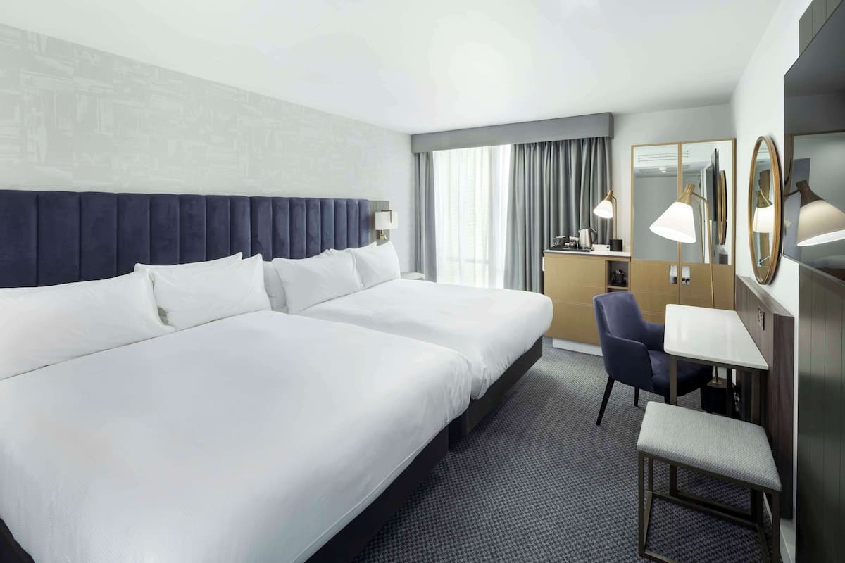 Ein Hotelzimmer mit zwei Betten, einem Schreibtisch, einem Stuhl, einer Bank, einem Spiegel und einem Fenster mit Vorhängen.