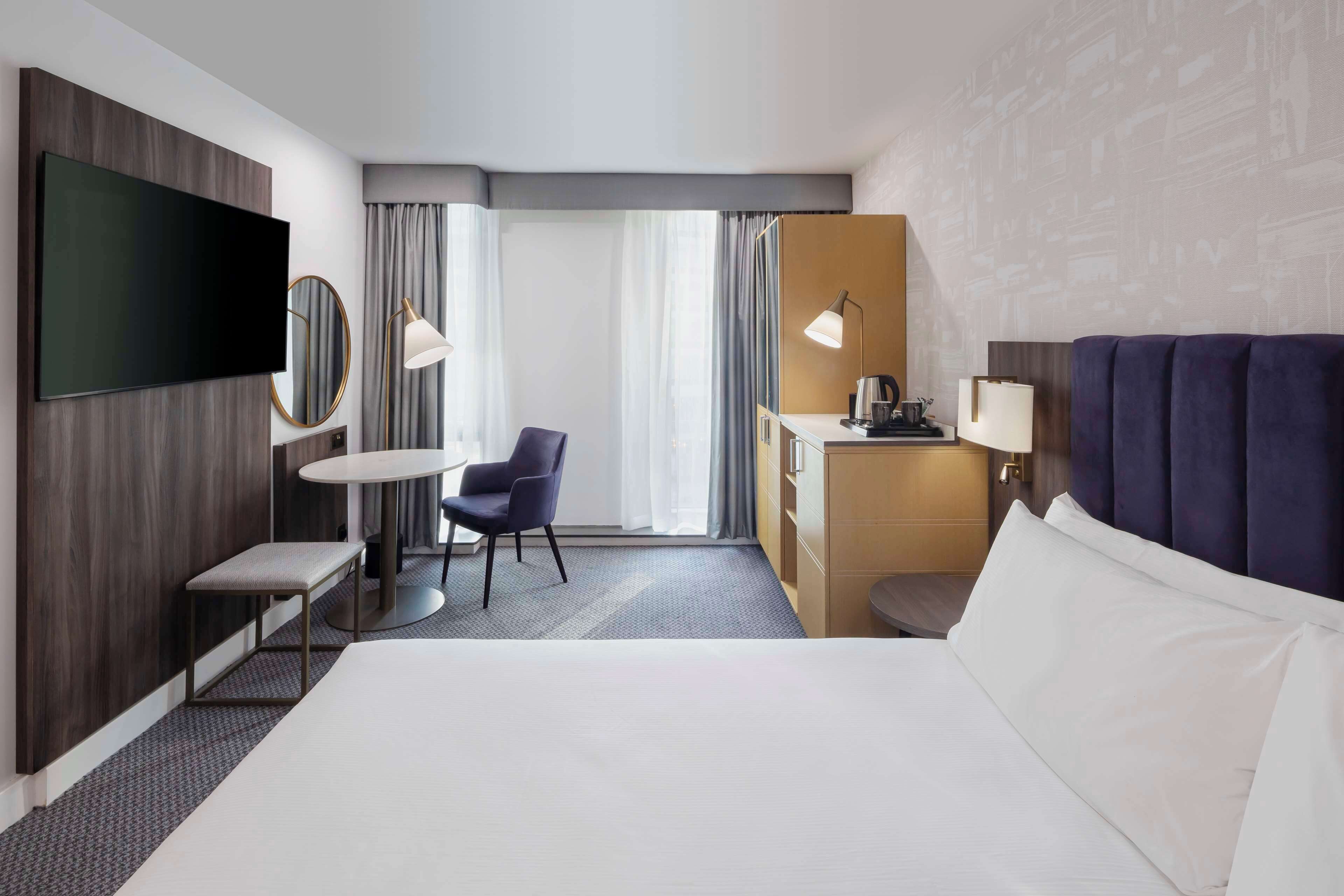 A modern hotel room with a large bed, a desk, a chair, a TV, and a window with curtains.