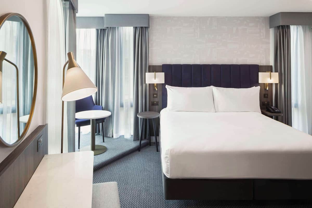 Ein Hotelzimmer mit einem großen Bett, zwei Nachttischen, einem Schreibtisch, einem Stuhl, einem runden Spiegel und einem Fenster mit Vorhängen.