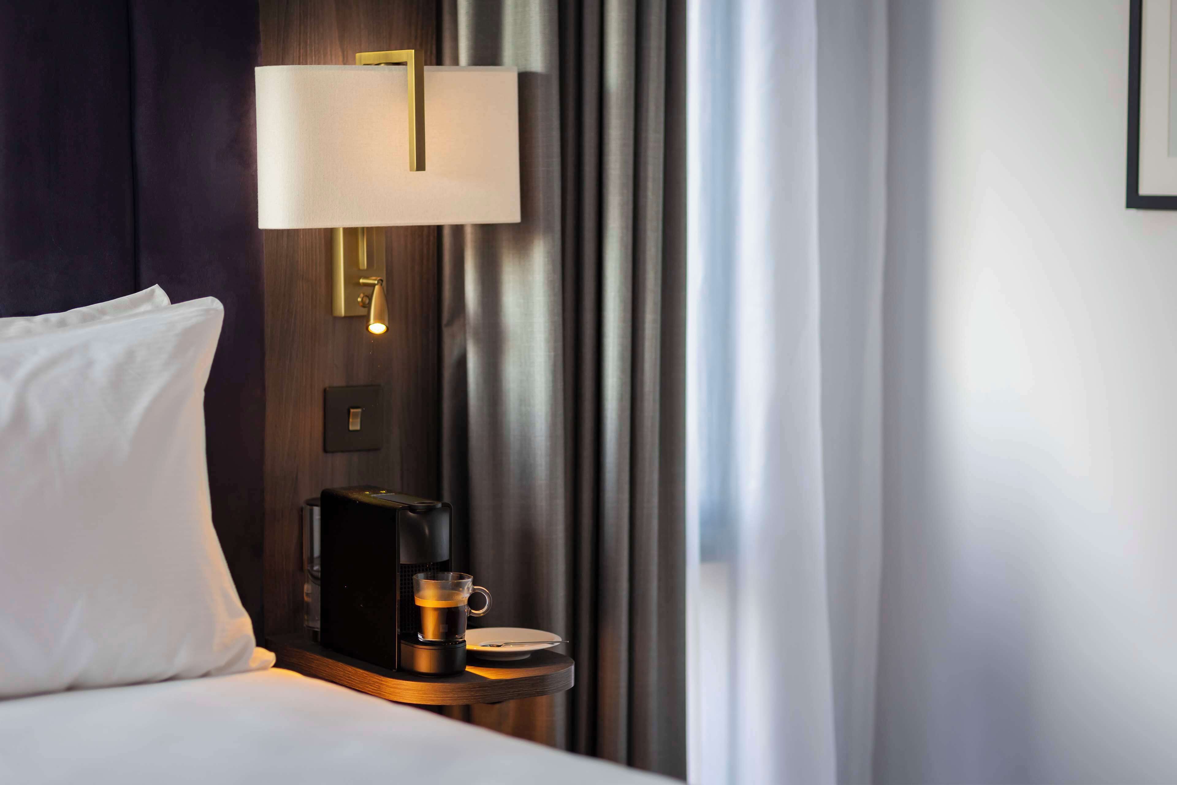 Een hotelkamer met een bed, een nachtkastje met een koffiezetapparaat, een lamp en een gordijn.