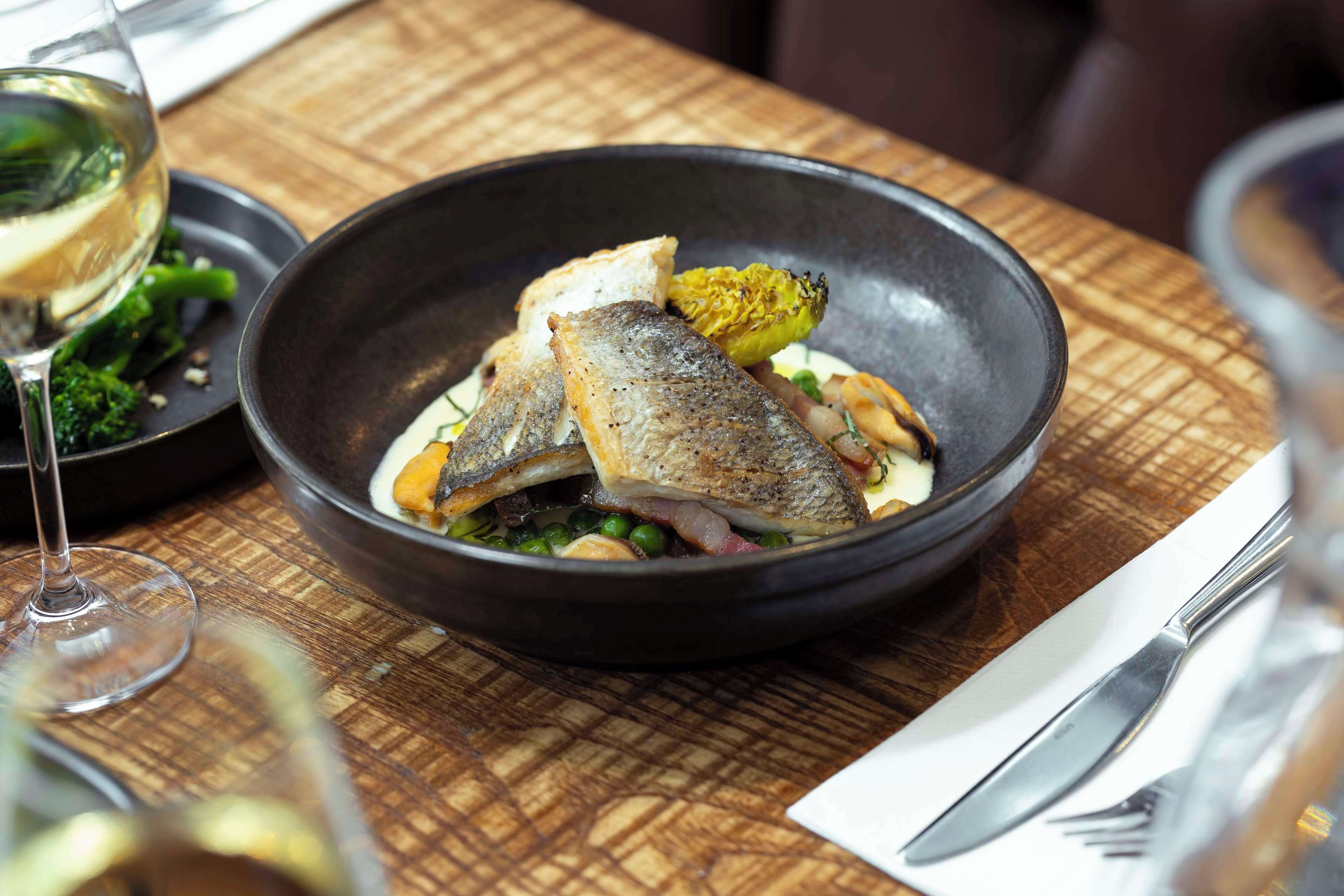 Une table à manger avec un bol noir contenant du poisson, des petits pois et un légume jaune, accompagné d’un verre de vin blanc.