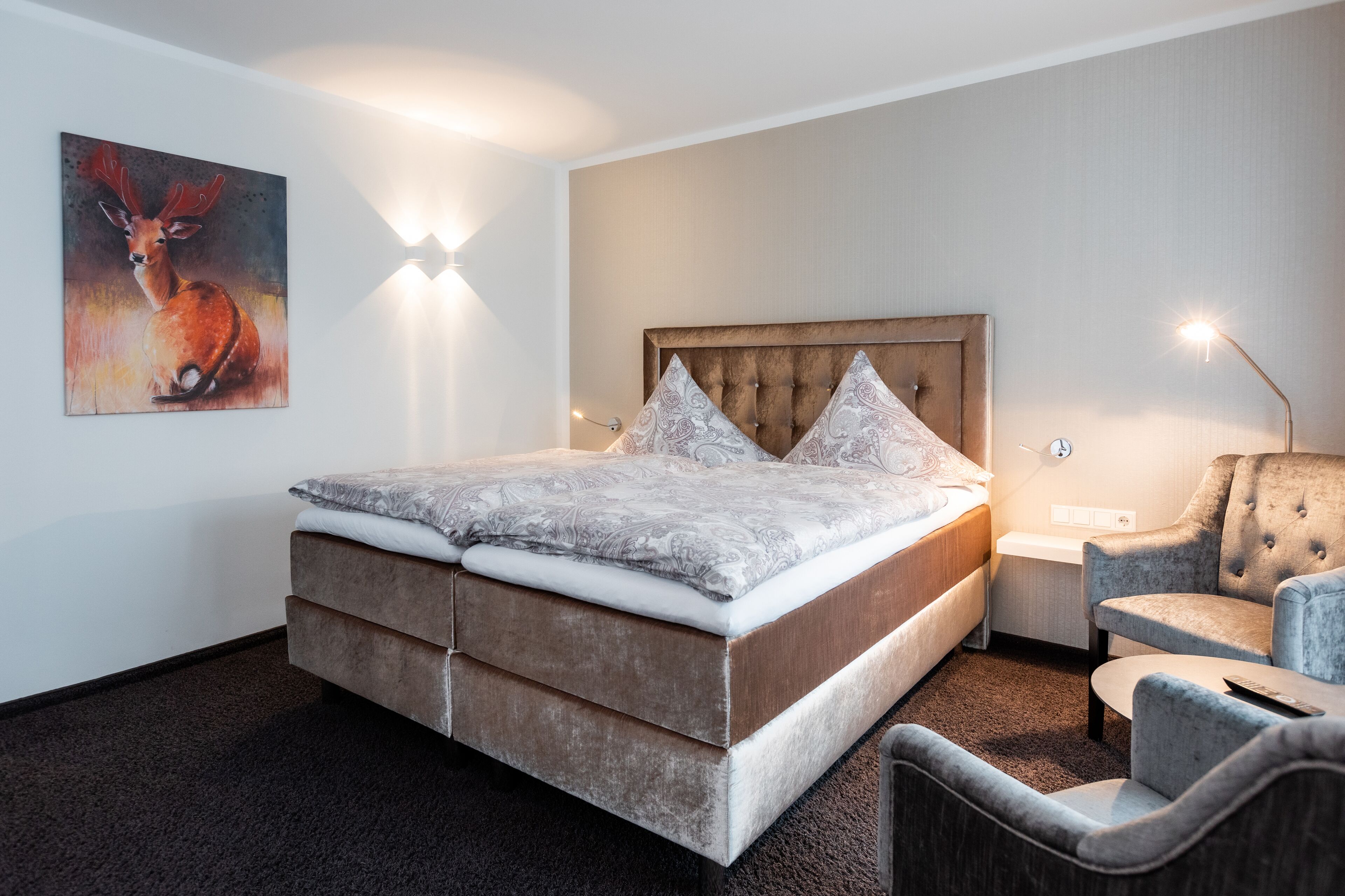 Un dormitorio moderno con una cama grande, un cabecero capitonado, una colcha gris estampada, un sillón gris y una lámpara fijada a la pared.