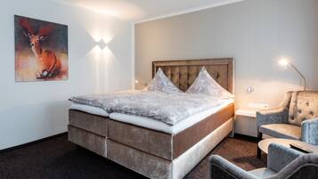 A modern bedroom with a large bed, a tufted headboard, a grey patterned bedspread, a grey armchair, and a wall-mounted light fixture.