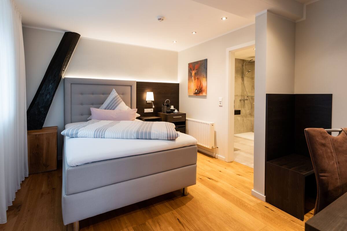 A modern bedroom with a large bed, bedside tables, a nightstand, a lamp, a painting, a shower area, and a wooden floor.