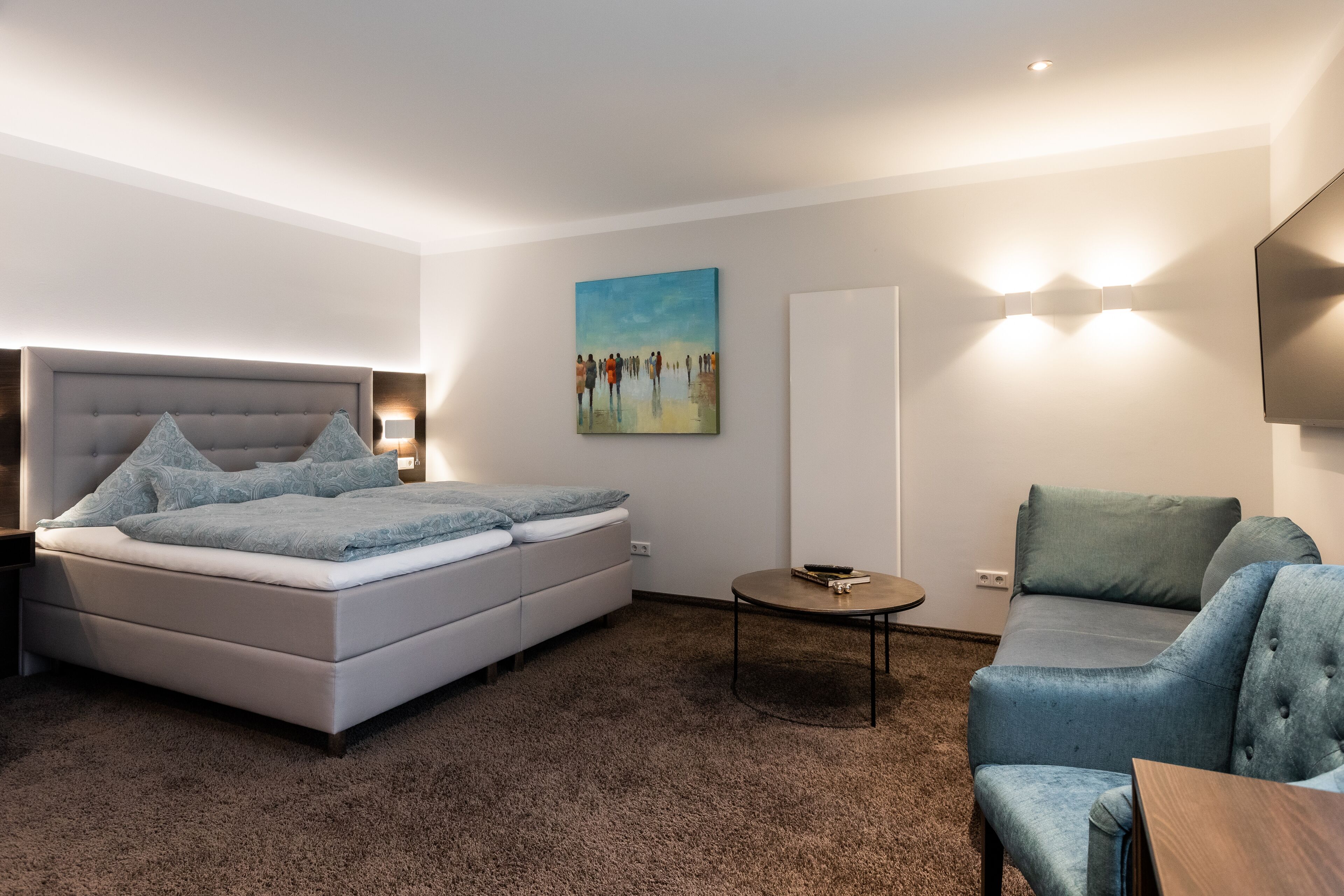 A modern hotel room with a large bed, a sofa, a small table, and a painting on the wall.