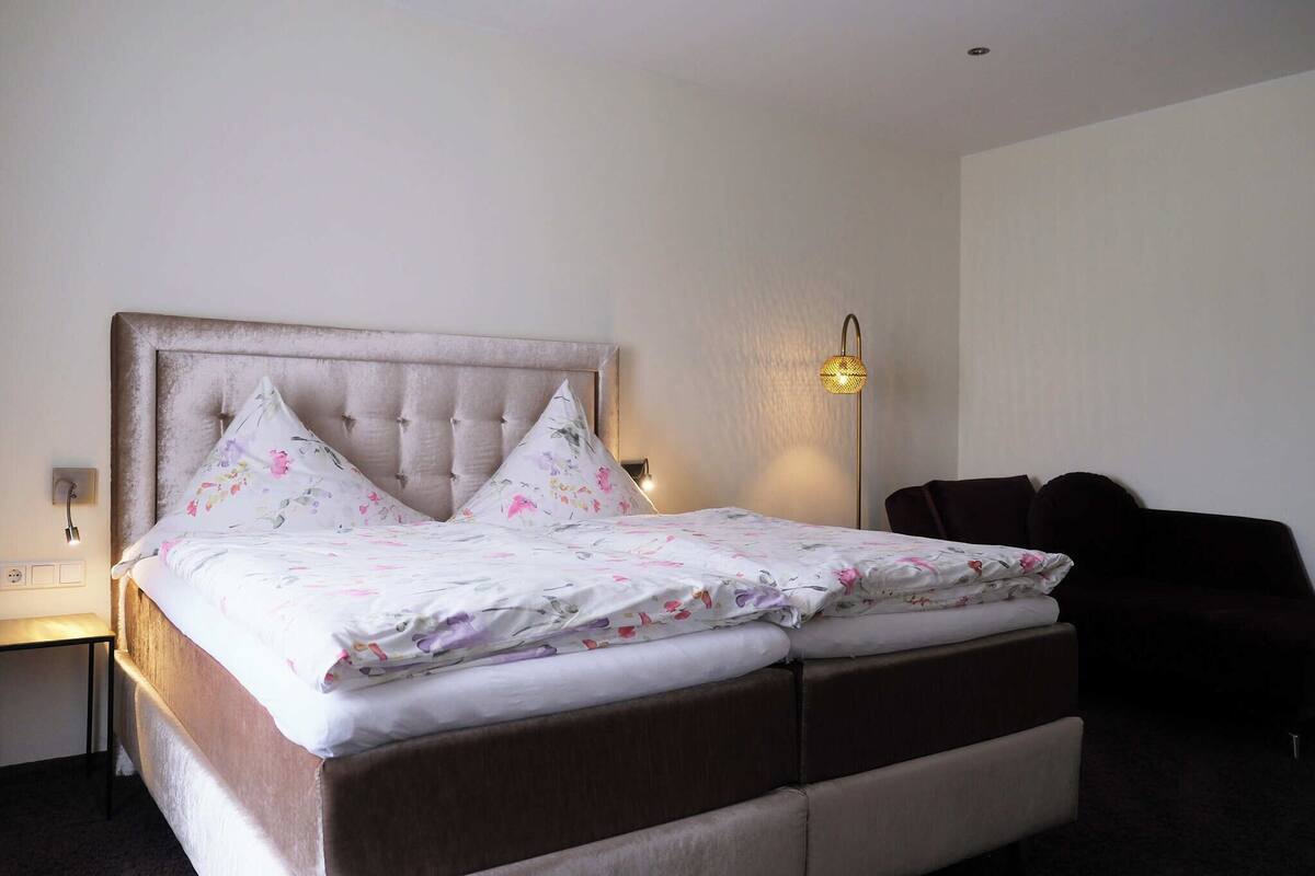 A neatly made bed with a tufted headboard, two pillows, and a floral-patterned bedspread. There is a bedside table with a lamp, and a dark-colored armchair to the right.