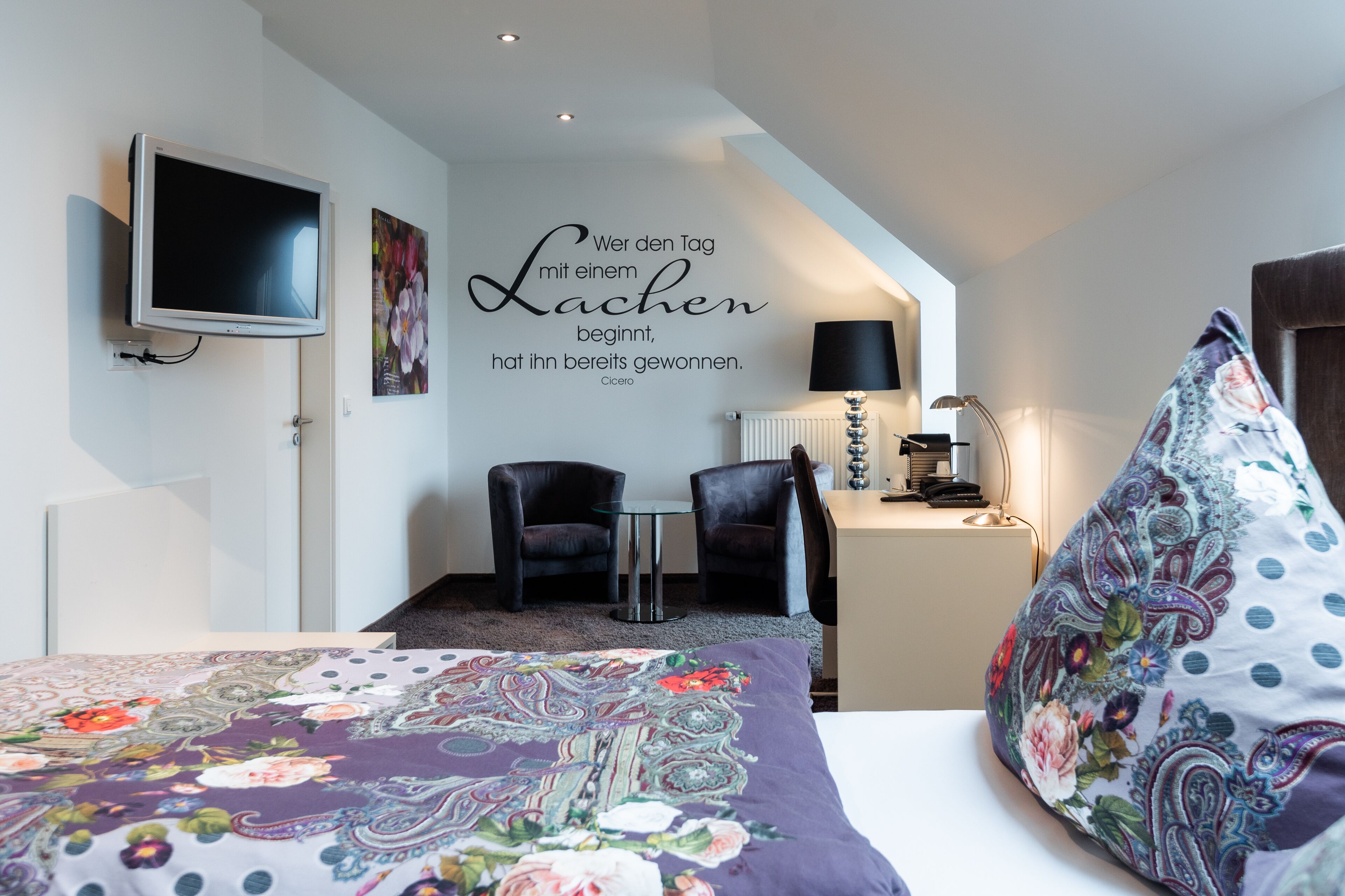 Habitación de hotel con cama, televisor montado en la pared, dos sillones, escritorio con lámpara y una cita decorativa en la pared.