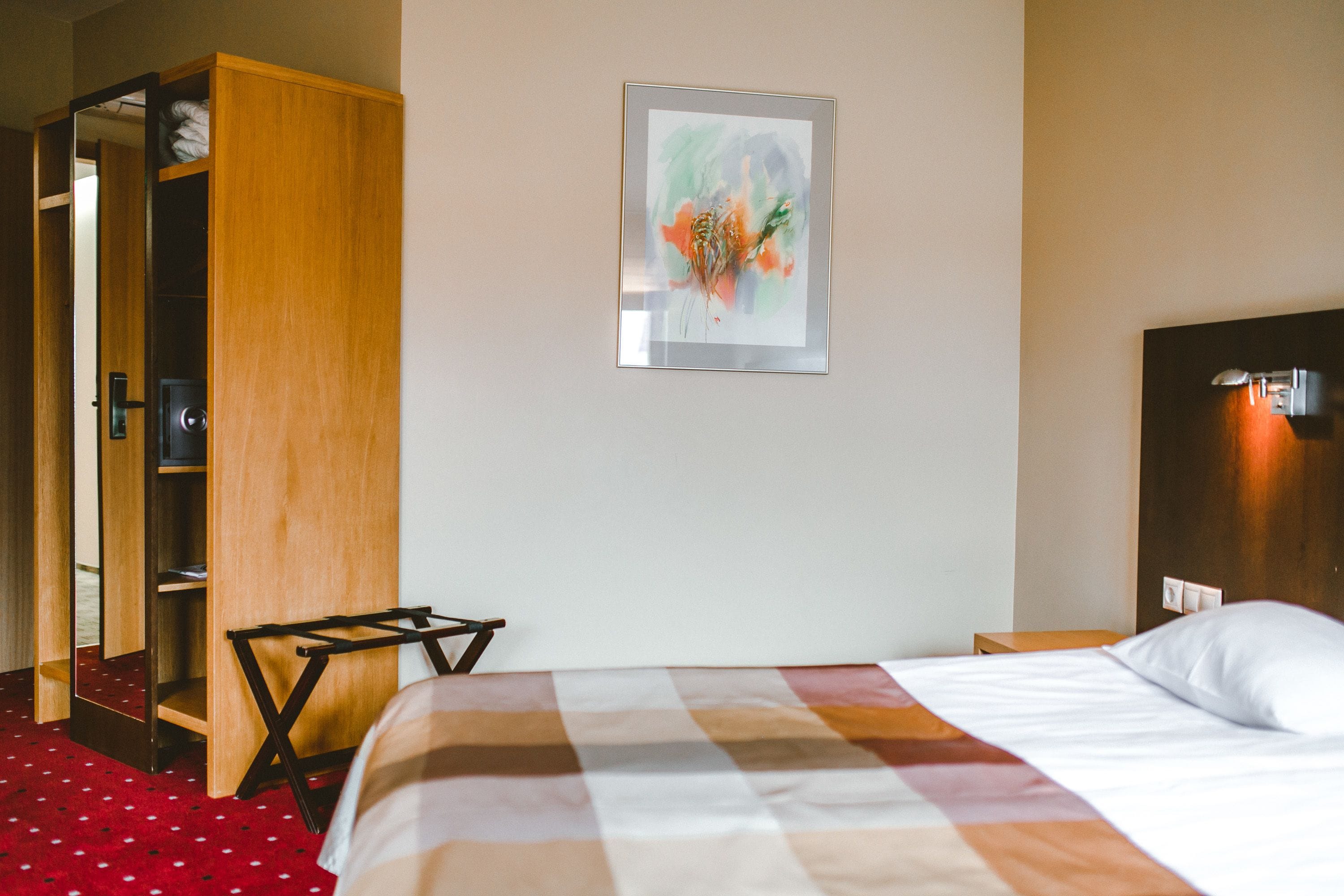 Quarto de hotel com uma cama, guarda-roupa de madeira, uma obra de arte emoldurada na parede e uma mesinha.
