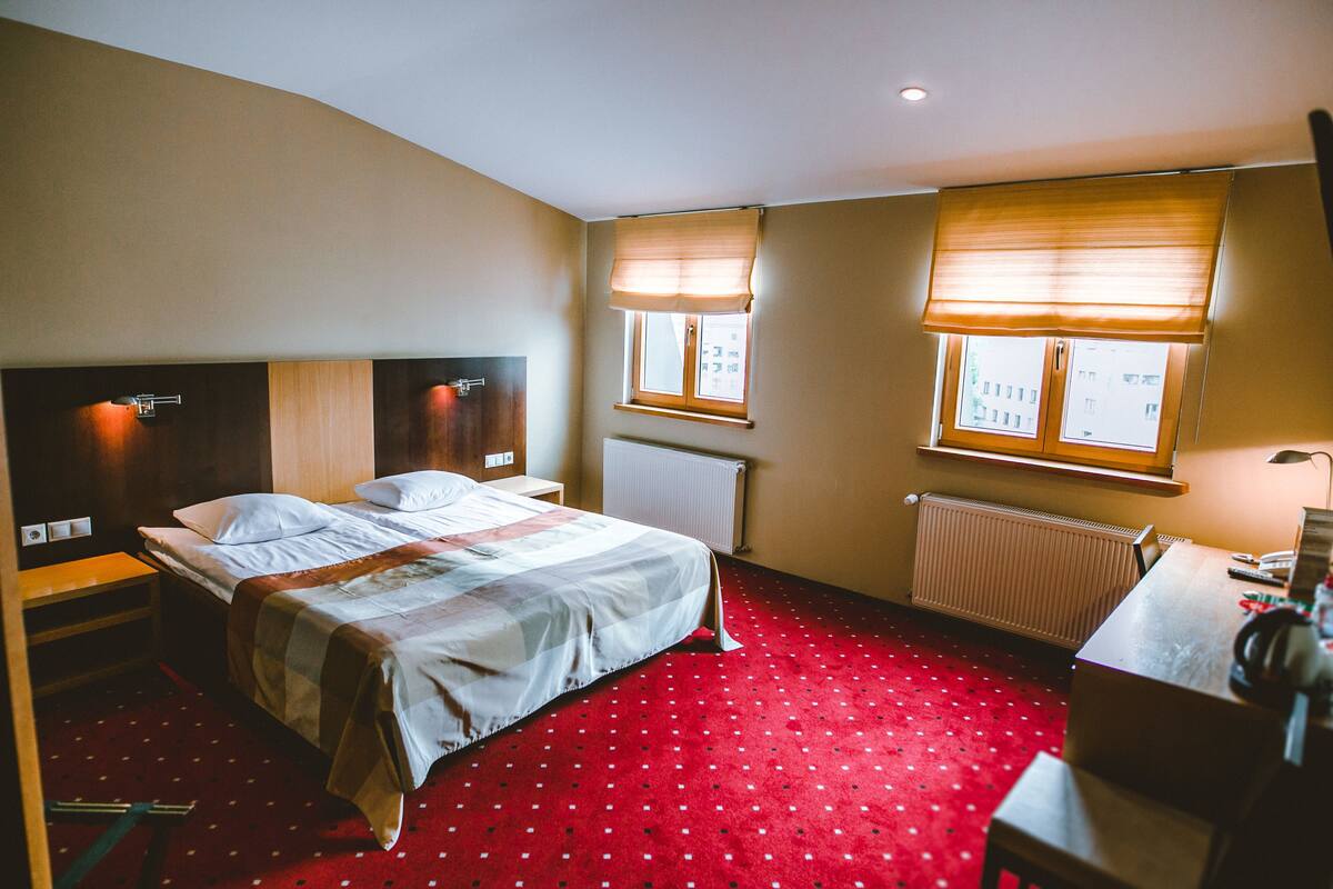 Una habitación de hotel con una cama, un escritorio, una silla y dos ventanas con persianas.