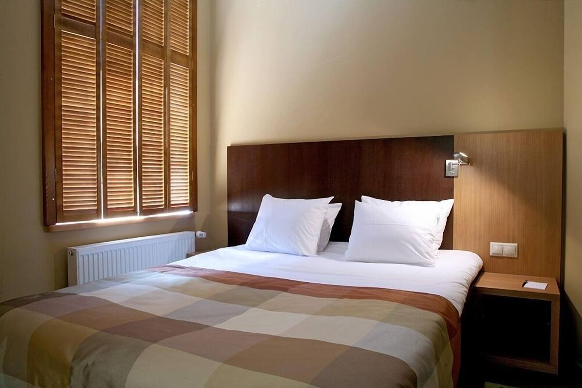 Habitación de hotel con una cama grande, cabecera de madera, una mesita de noche y una ventana con persianas.