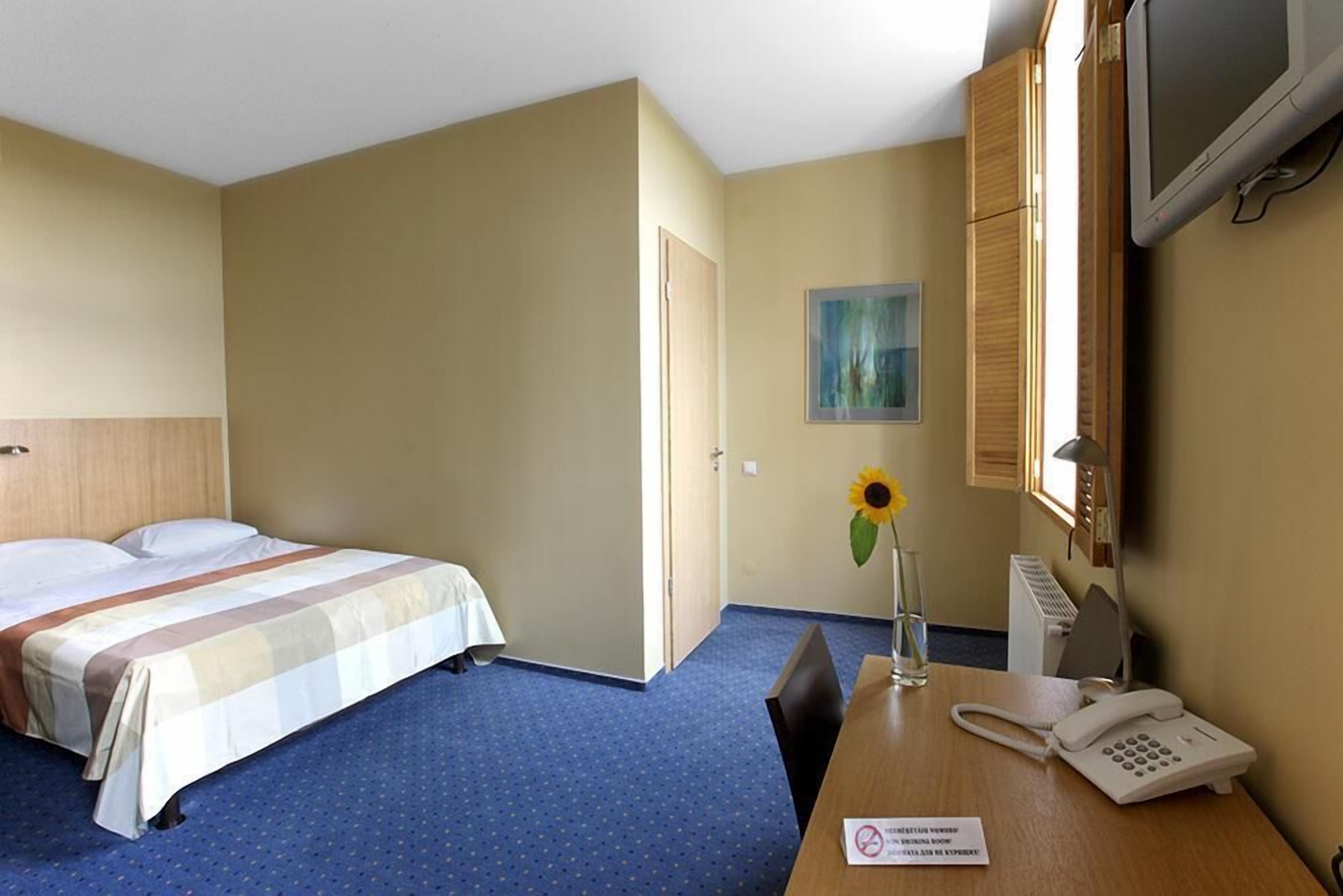 Hotellihuone, jossa on sänky, kirjoituspöytä puhelimineen, tuoli, pieni pöytä, taulu seinällä ja ikkuna.