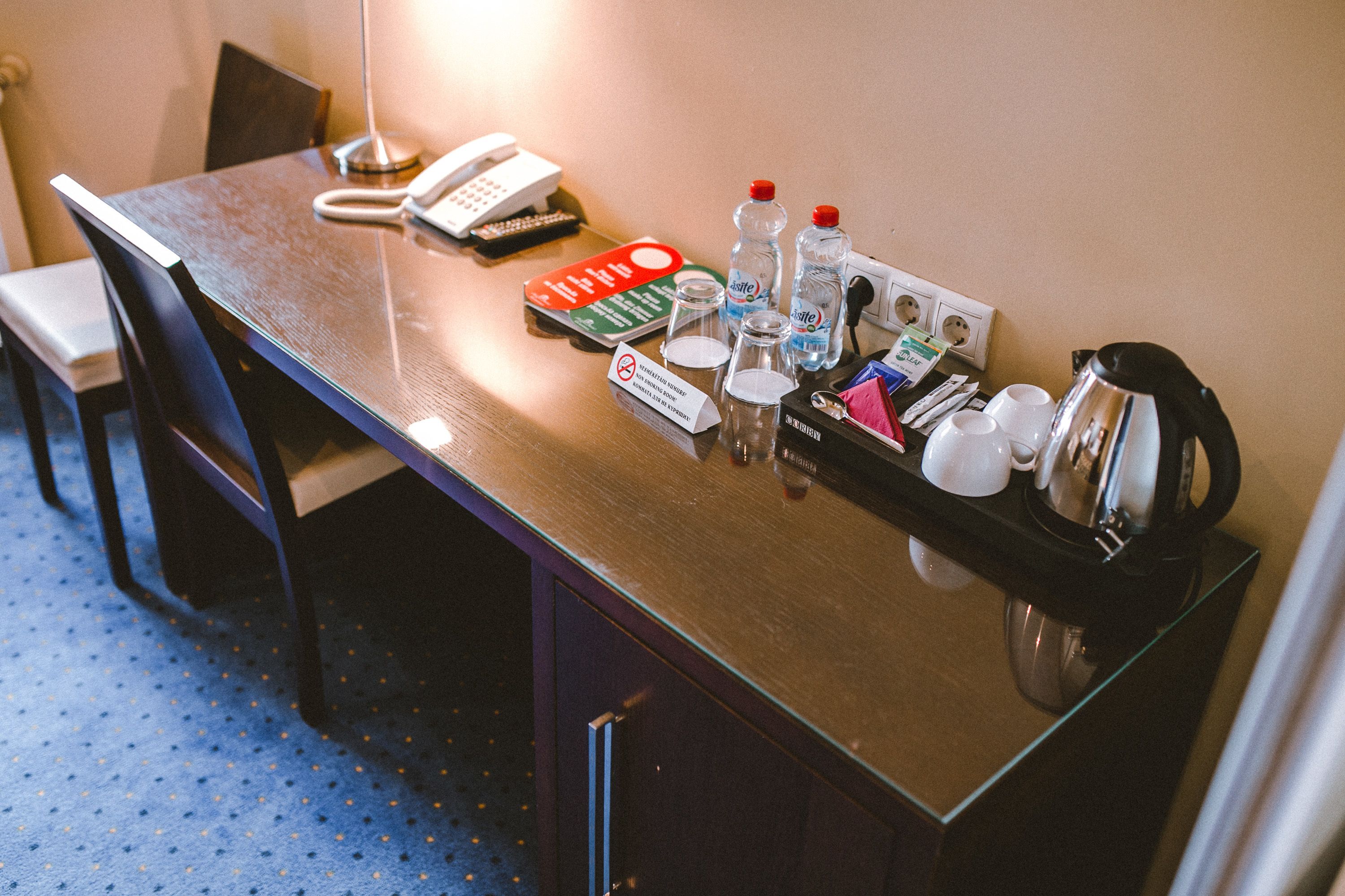 A hotel room desk with a telephone, a notepad, a pen, a glass of water, a bottle of water, a coffee pot, and a small box of tissues.