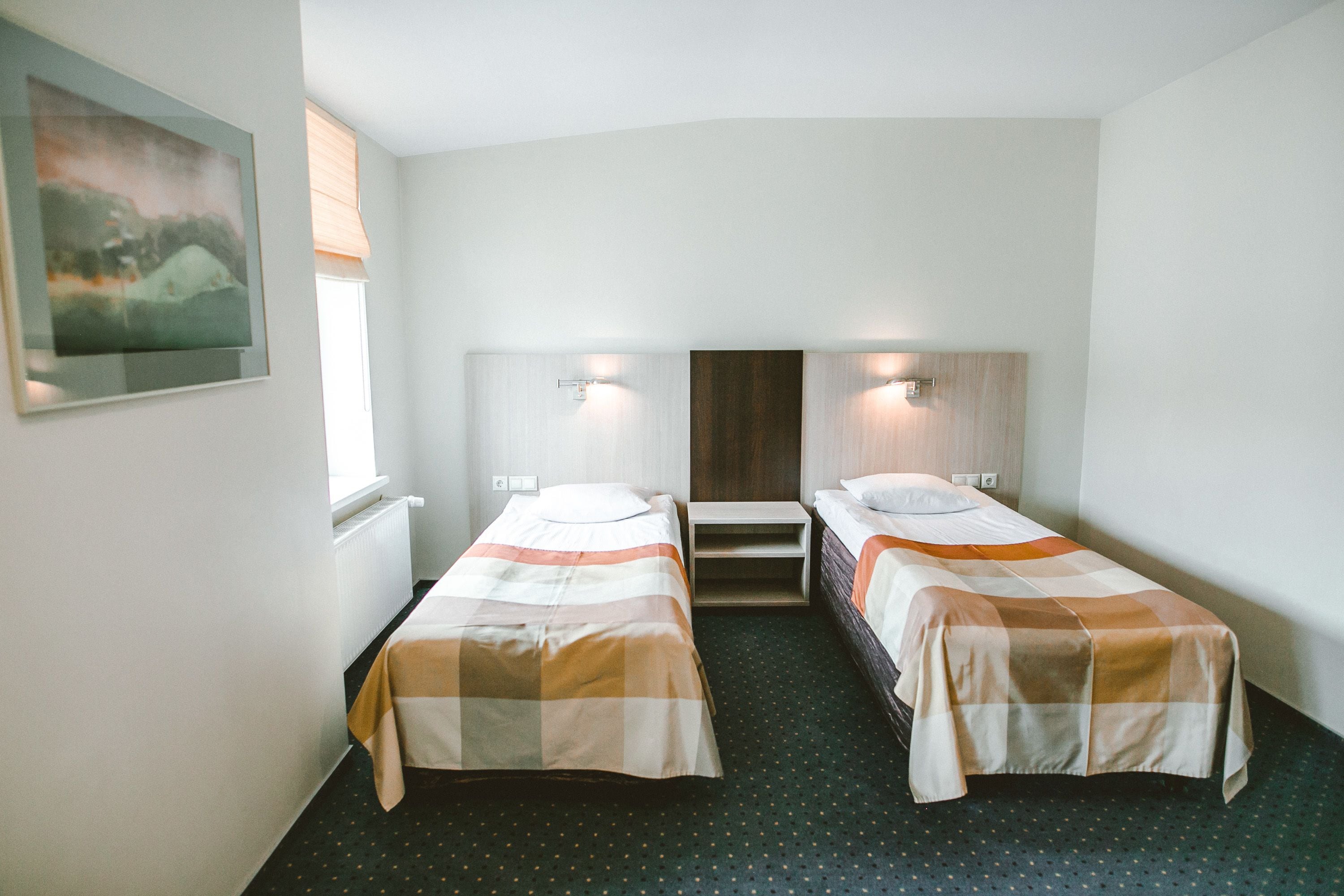 Hotellihuone, jossa on kaksi erillistä sänkyä, pieni pöytä, ikkuna, jossa on sälekaihtimet, ja taulu seinällä.