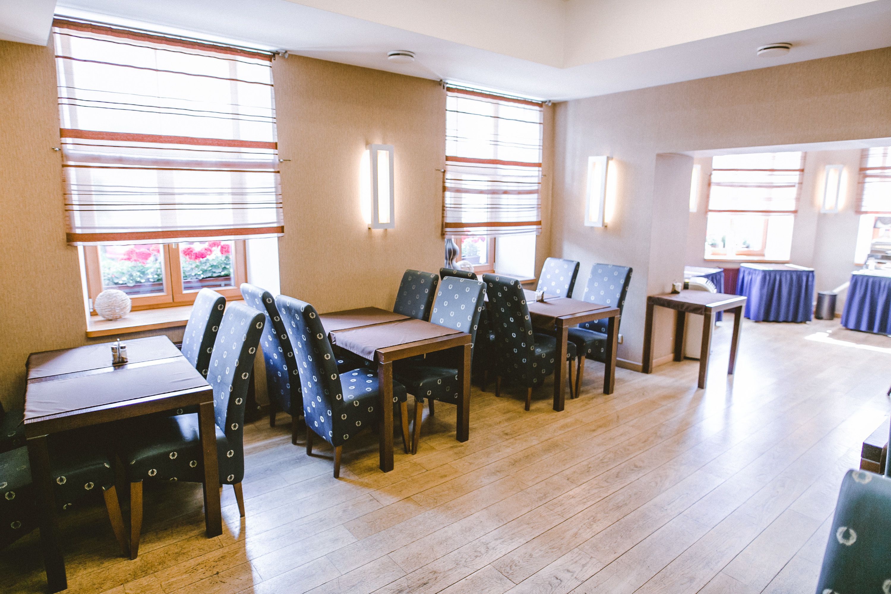 A dining area with wooden tables and chairs, large windows with blinds, and a view of the outdoors.