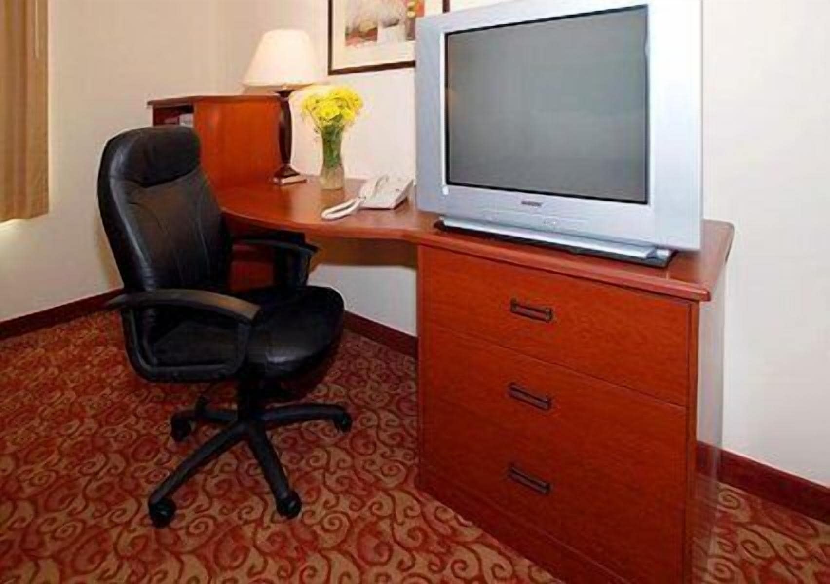 Quarto de hotel com secretária de madeira, cadeira de escritório preta, televisão de tela plana e telefone.