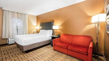Habitación de hotel con cama, sofá rojo, dos lámparas de noche, ventana con cortinas y alfombra estampada.