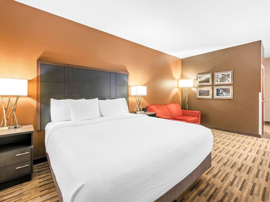 Hotellihuone, jossa on suuri sänky, kaksi yöpöytää, yölamppu, punainen tuoli ja seinälle asennettu televisio.