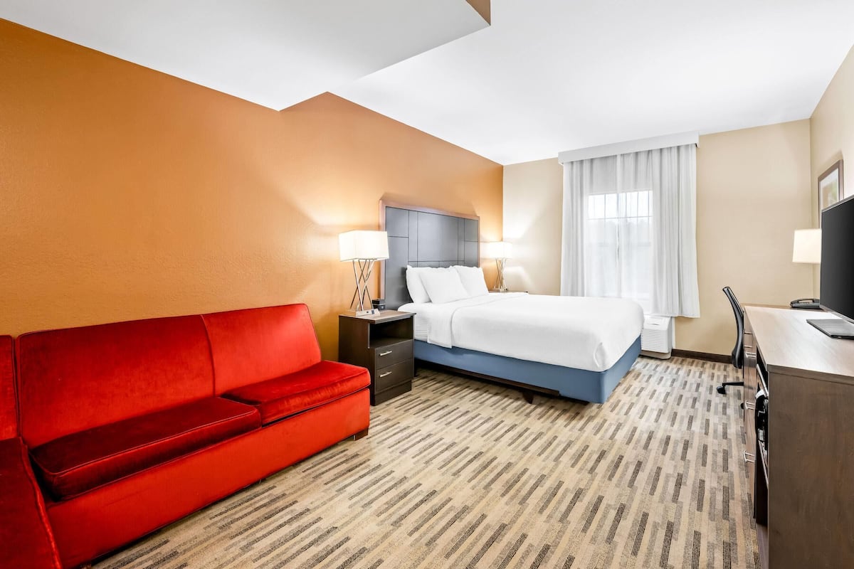 Habitación de hotel con un sofá rojo, una cama con ropa de cama blanca, un escritorio con una computadora y un televisor.
