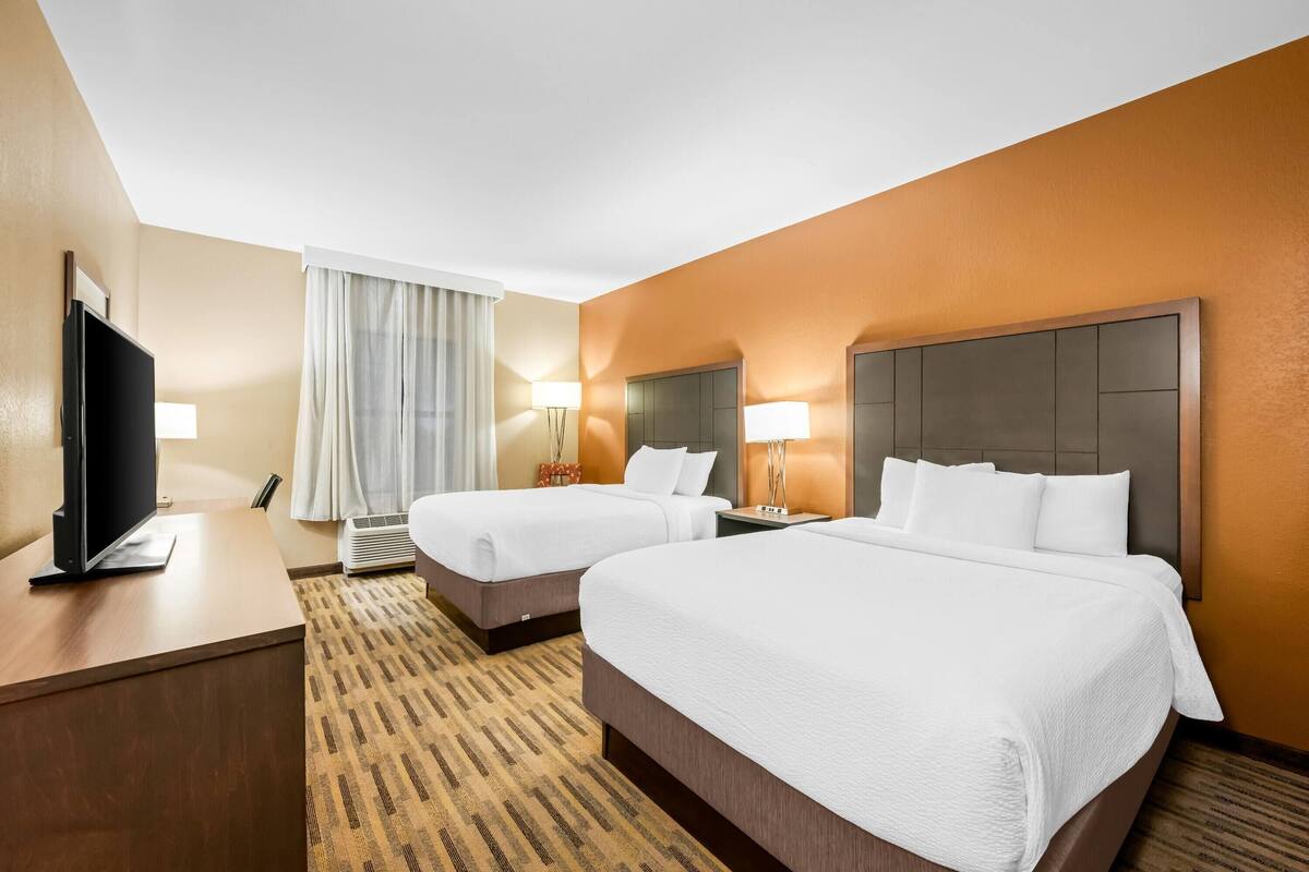 A hotel room with two beds, a desk, a TV, and a large window with curtains.