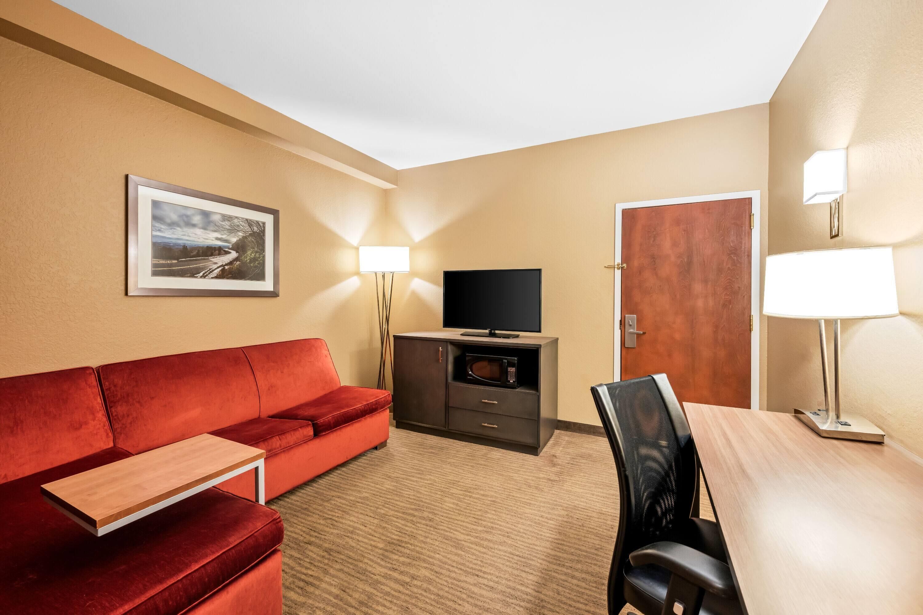A hotel room with a red sofa, a wooden table, a TV on a cabinet, a door, and a lamp.