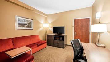 Una habitación de hotel con un sofá rojo, una mesa de madera, un televisor sobre un mueble, una puerta y una lámpara.