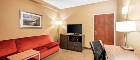A hotel room with a red sofa, a wooden table, a TV on a cabinet, a door, and a lamp.