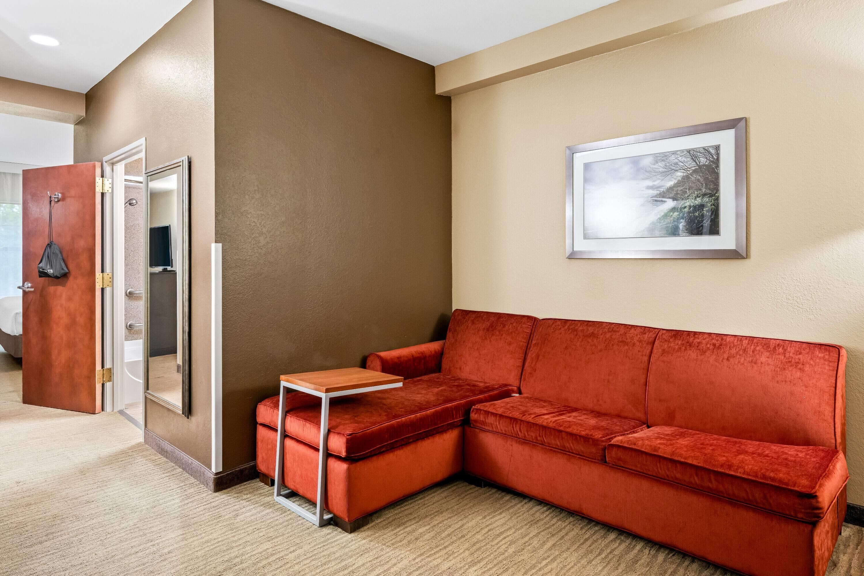 Una habitación de hotel moderna con un sofá de terciopelo rojo, una mesita de madera pequeña y un cuadro en la pared.