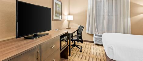 A hotel room with a flat-screen TV, a desk with a chair, a bed, a lamp, and a window with curtains.