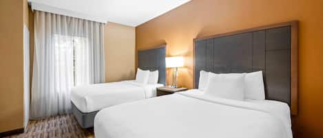 A hotel room with two beds, a large window with sheer curtains, a bedside table with a lamp, and a patterned carpet.