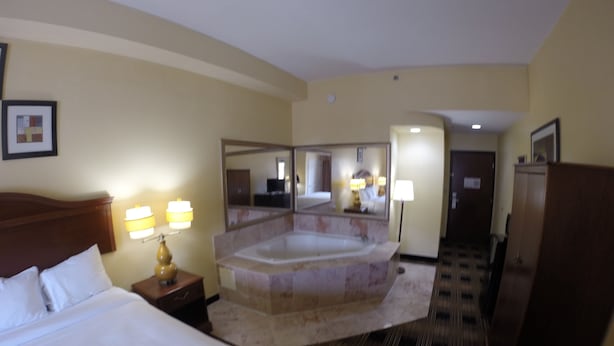 A hotel room with a large bed, a sitting area with a chair and a table, a large bathtub, and a mirror. - Jameson Inn and Suites Riverdale (Riverdale)