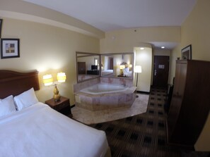 A hotel room with a large bed, a sitting area with a chair and a table, a large bathtub, and a mirror. - Jameson Inn and Suites Riverdale (Riverdale)