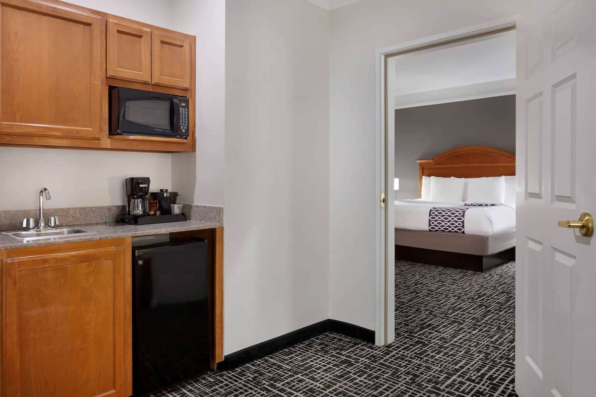 A compact hotel room with a kitchenette including a microwave, coffee maker, and sink. A bed with a headboard and a patterned carpet is visible through an open door.