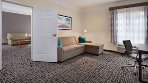 Habitación de hotel con sofá color beige, una mesita, un escritorio con silla, alfombra estampada, una puerta blanca y una ventana con cortinas.
