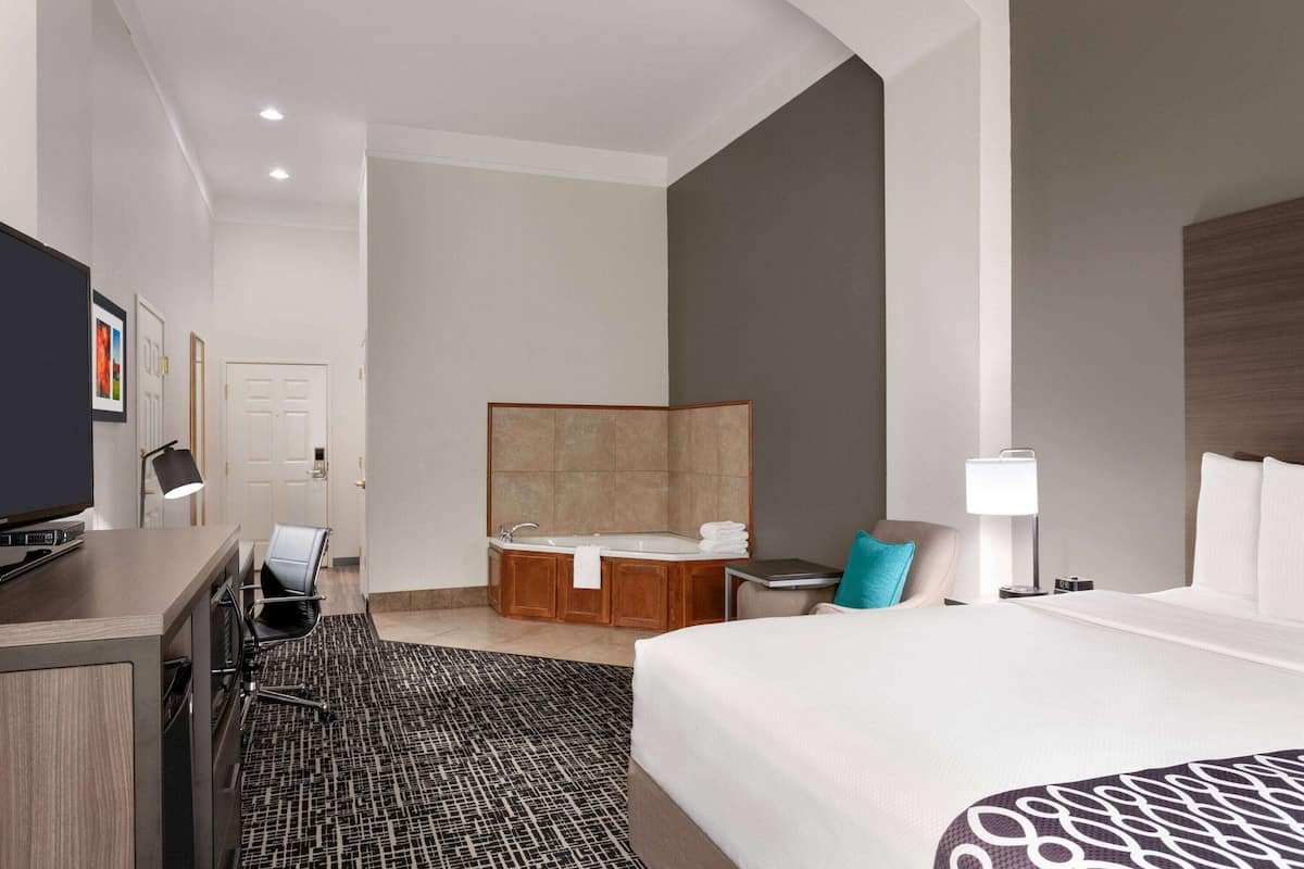 A hotel room with a large bed, a desk with a computer, a chair, a bathroom with a bathtub, and a TV.