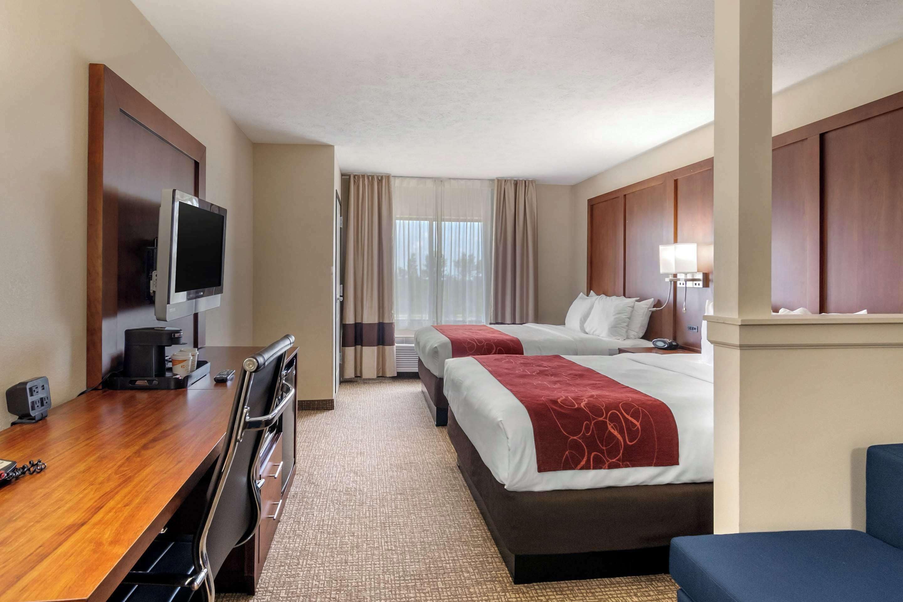 A hotel room with a wooden desk, two beds, a TV, and a window with curtains.