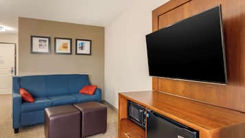 Habitación de hotel con un sofá azul, dos otomanas marrones, un mueble de madera con microondas y un televisor de pantalla plana montado en la pared.