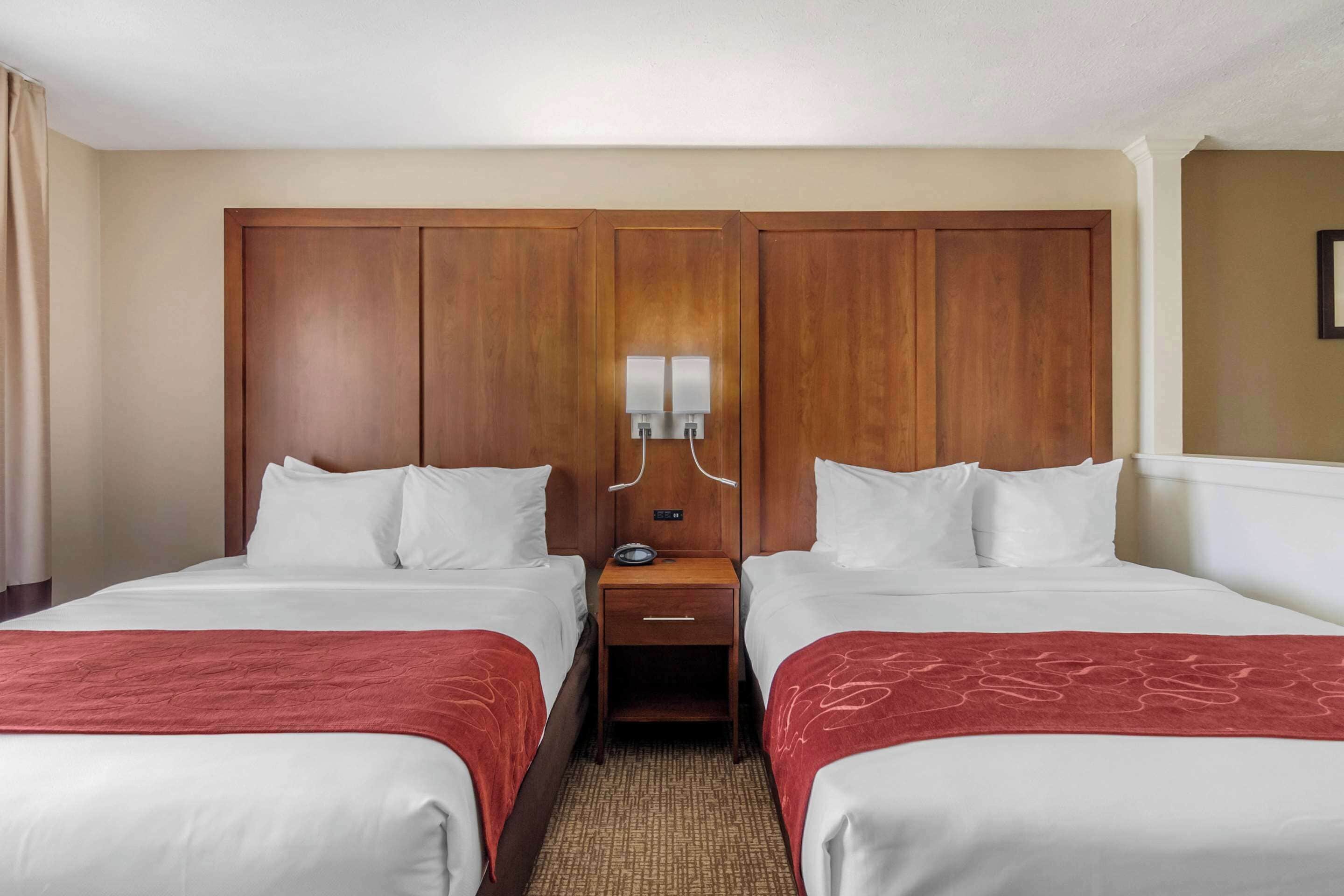 A hotel room with two beds, a wooden headboard, a nightstand with a lamp, and a red throw blanket.