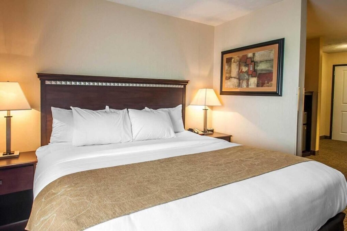 Een hotelkamer met een groot bed, twee nachtkastjes met lampen, een lijst met kunstwerk aan de muur, en een deur die naar een andere kamer leidt.