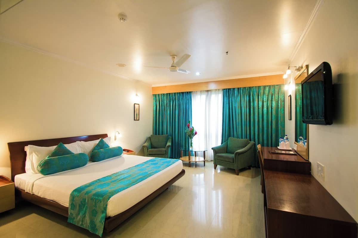 A hotel room with a bed, two armchairs, a small table, a TV, and a ceiling fan.