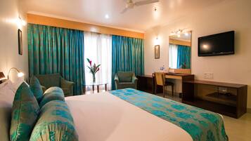 A hotel room with a bed, two chairs, a desk, a television, and a ceiling fan.