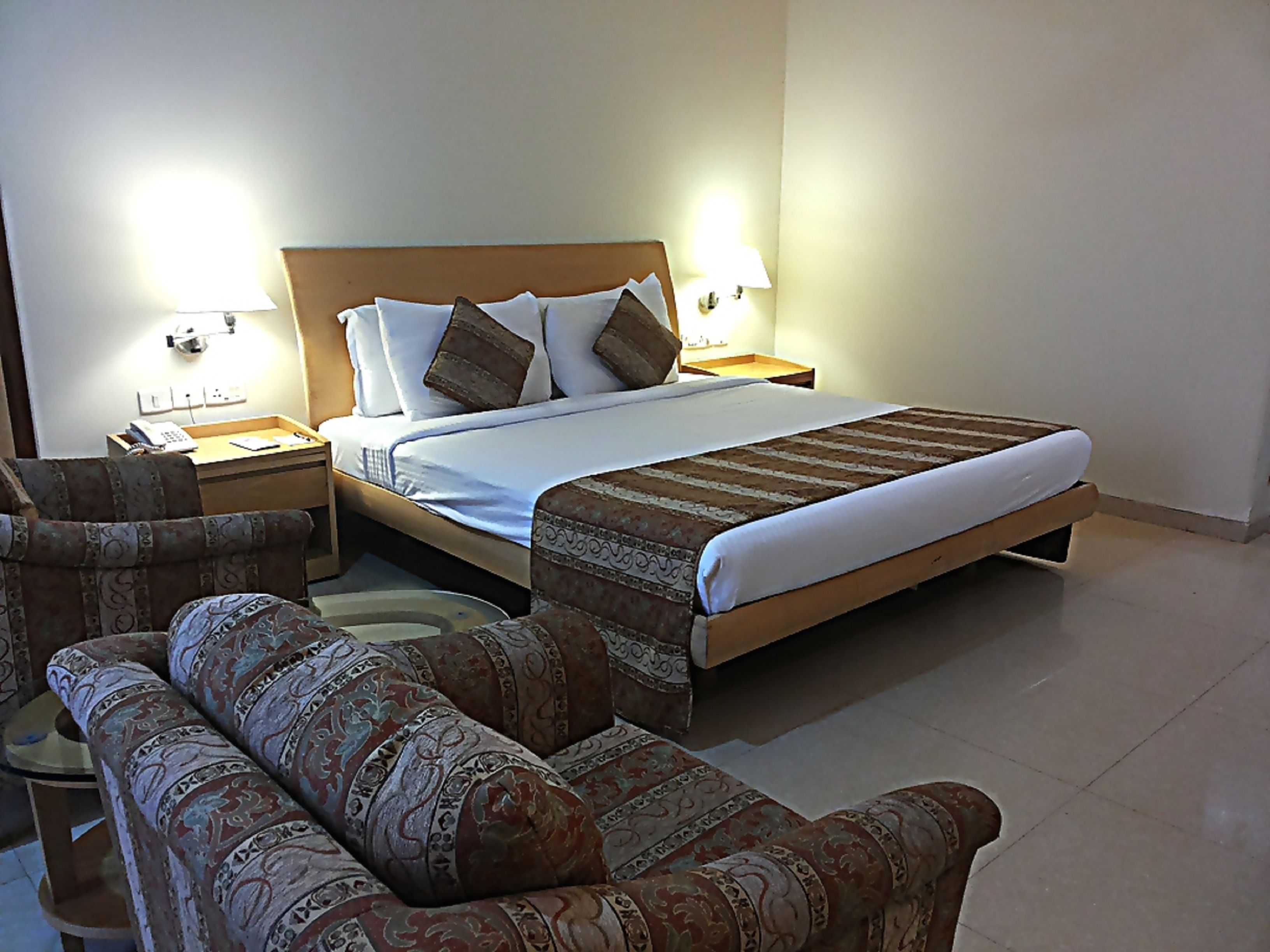 Una camera d'albergo con un letto, due comodini, una lampada, un divano con motivo decorativo e un piccolo tavolino rotondo.