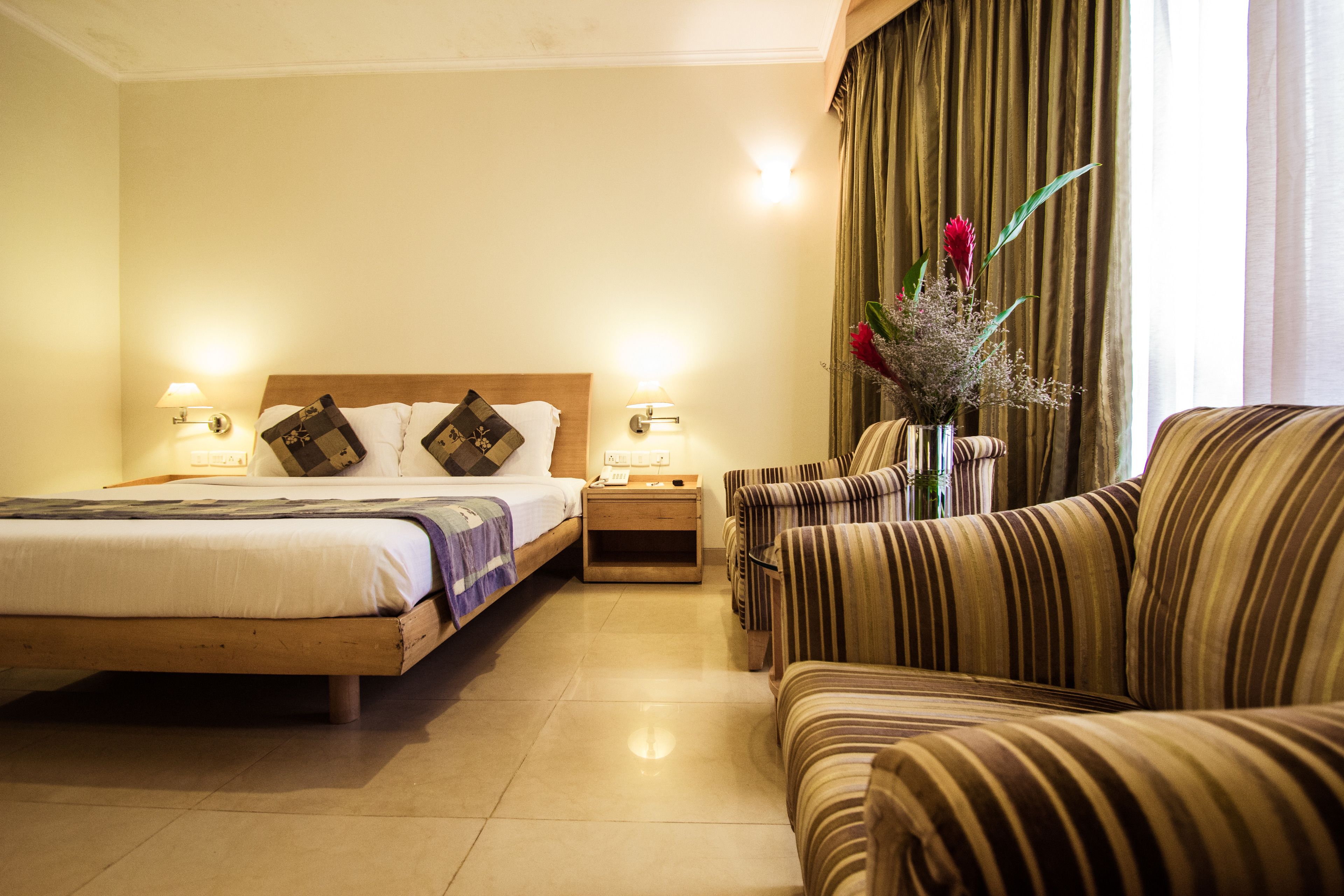 Habitación de hotel con una cama, dos mesitas de noche, un sofá a rayas y un florero con flores.