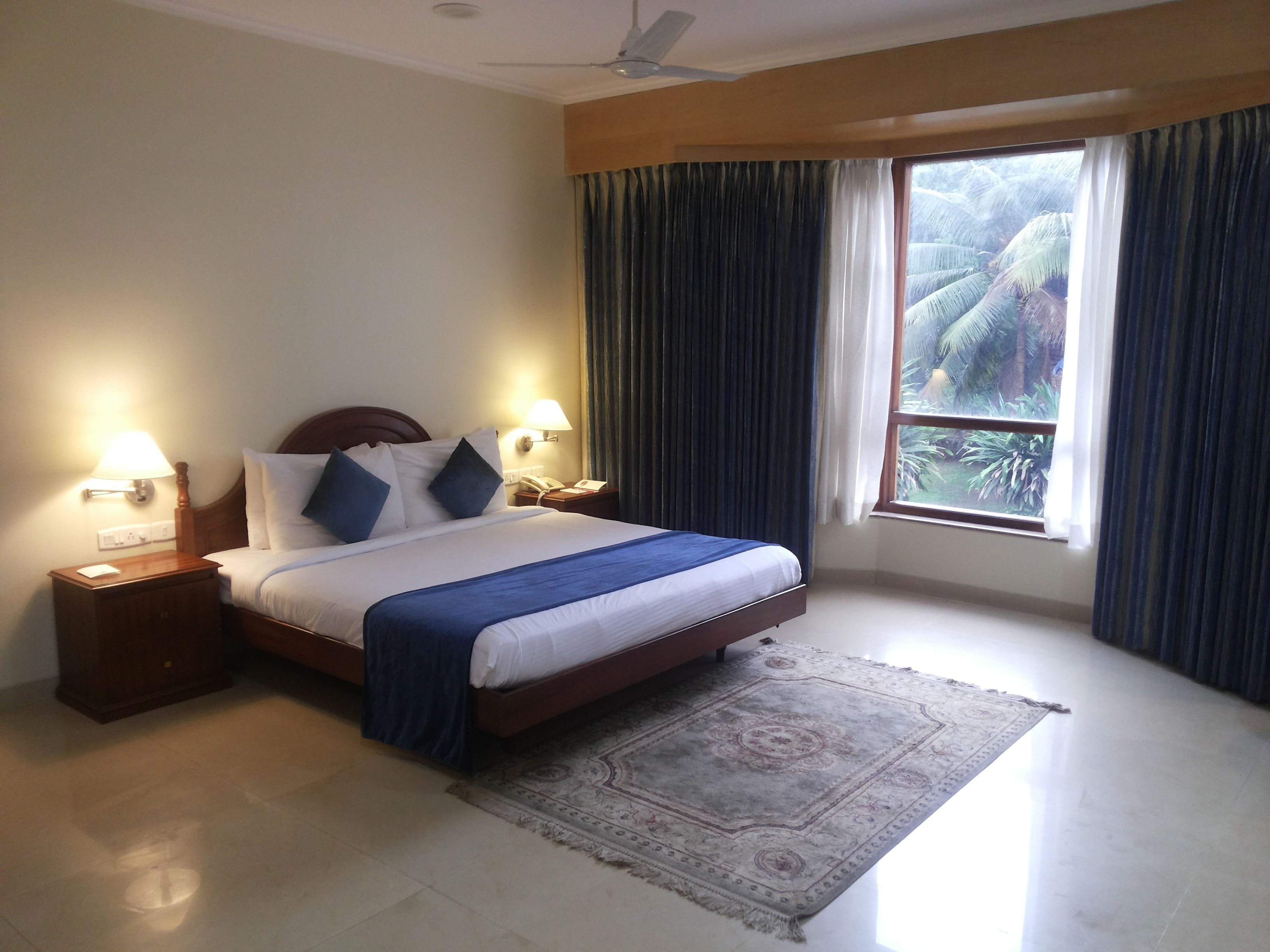 A hotel room with a bed, nightstands, lamps, a ceiling fan, a window with curtains, and a view of palm trees.