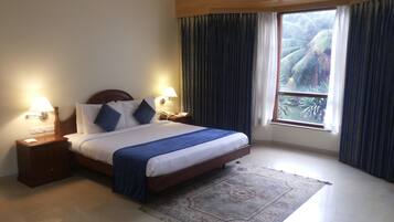 A hotel room with a bed, nightstands, lamps, a ceiling fan, a window with curtains, and a view of palm trees.