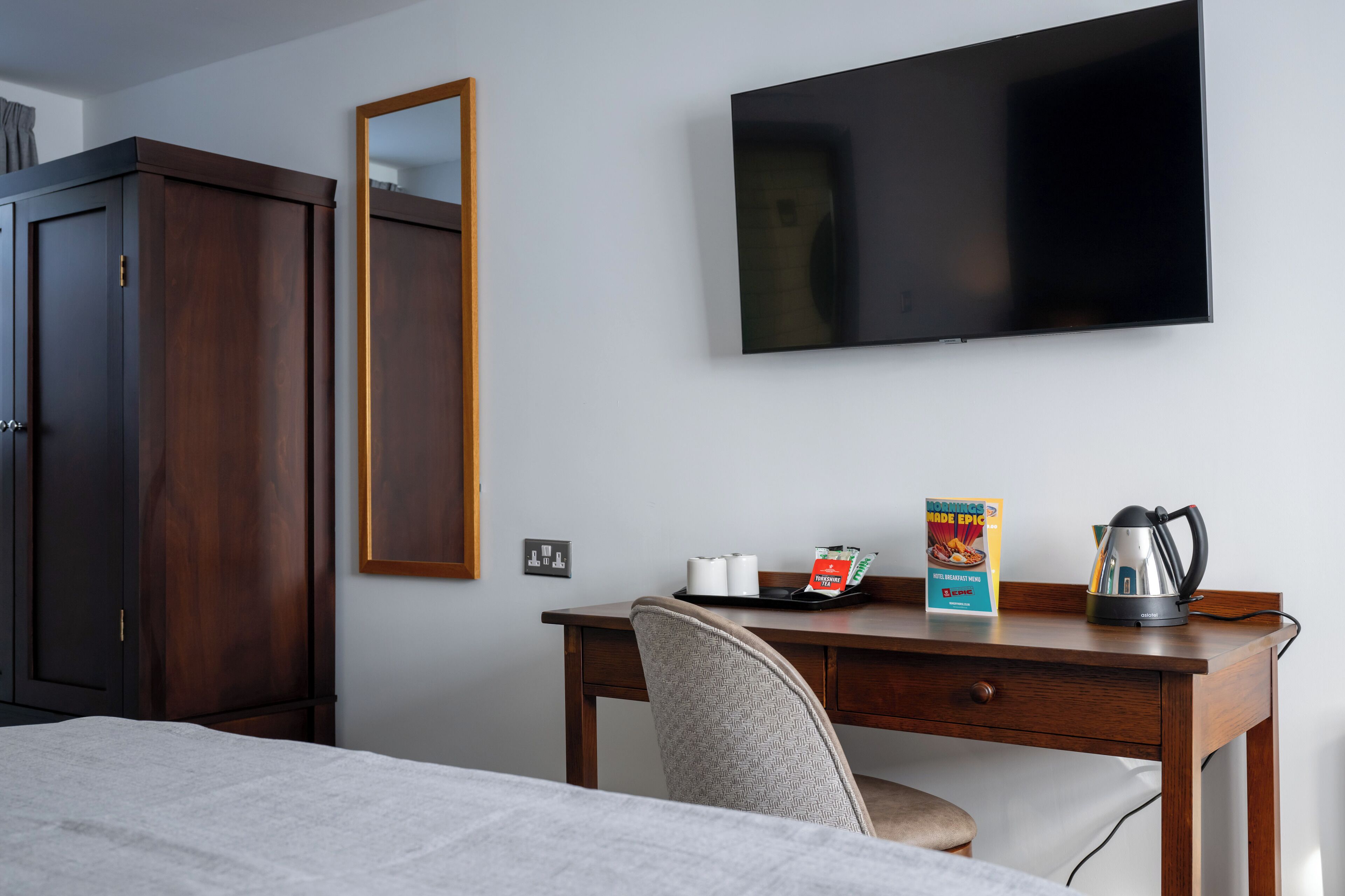A hotel room with a bed, a desk with a chair, a television, a wardrobe, and a mirror.