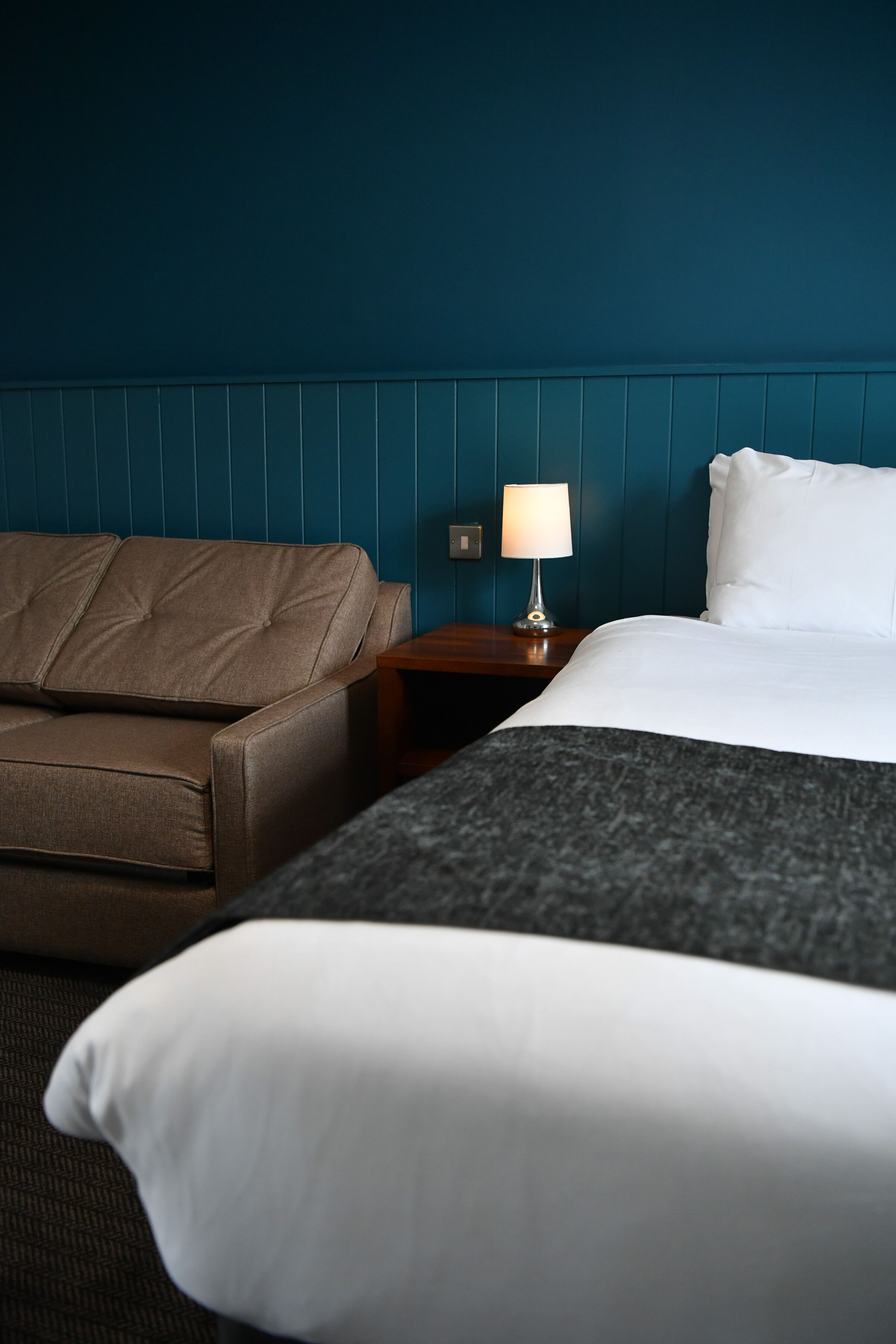 Quarto de hotel com uma cama, um sofá de couro marrom, uma mesa de cabeceira com luminária e uma parede azul.