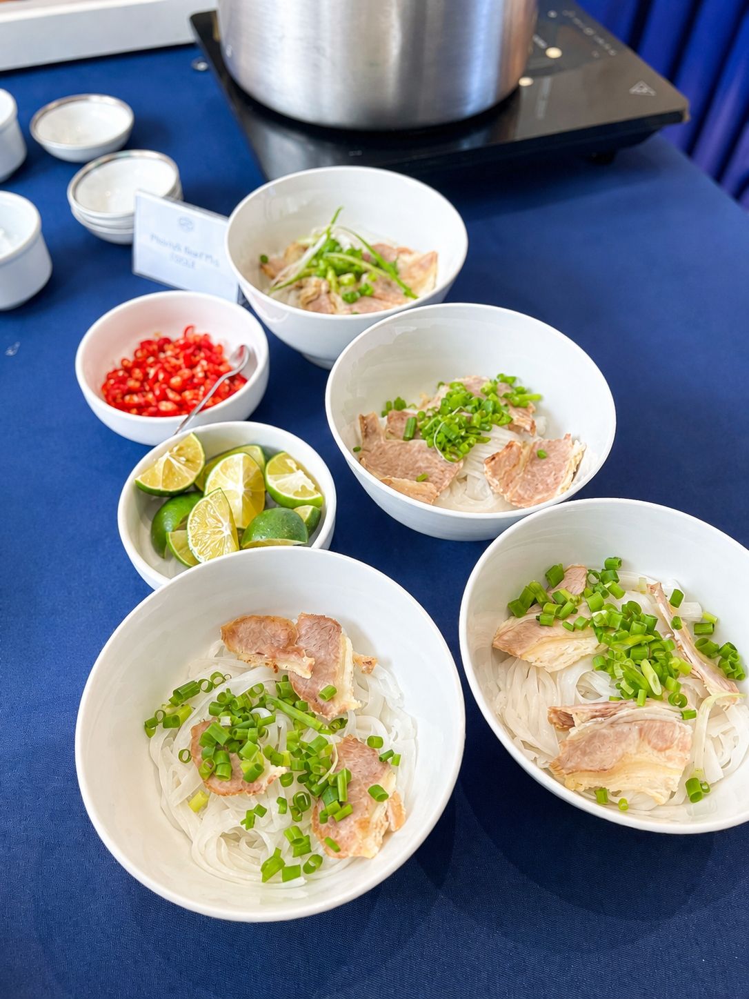 A table with bowls of noodle soup garnished with green onions and slices of meat, accompanied by small bowls of lime and chili peppers.