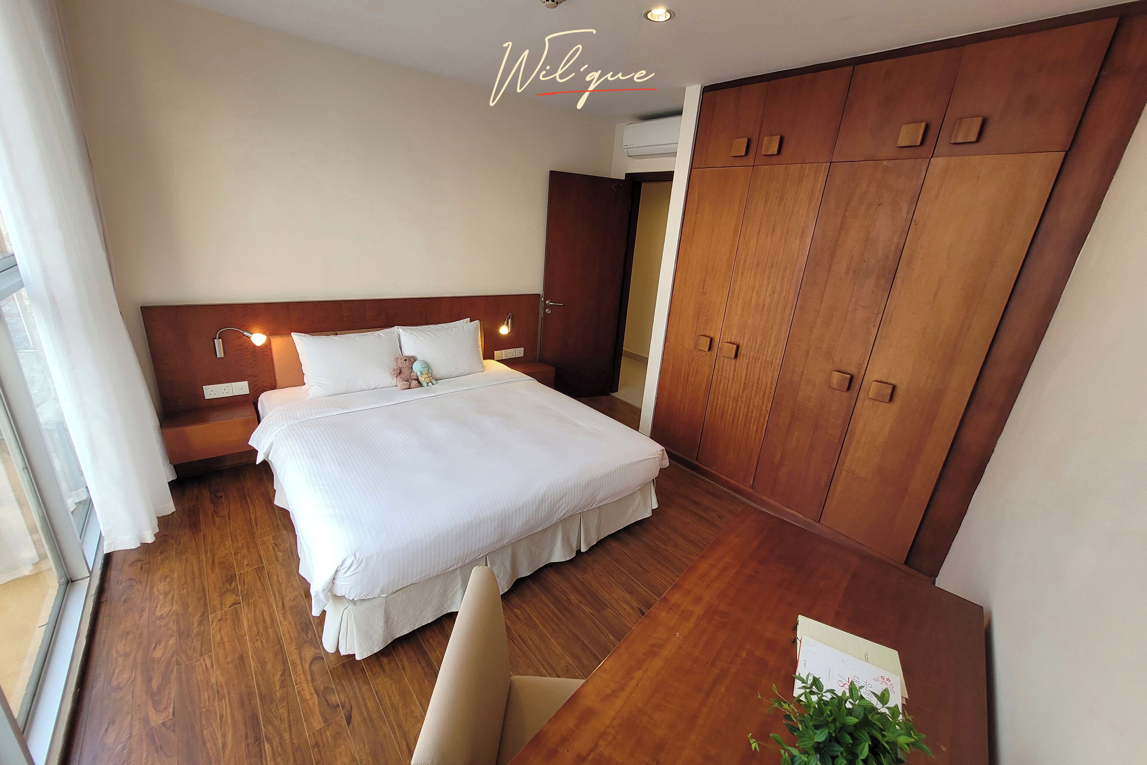 A hotel room with a large bed, wooden wardrobe, and a desk with a plant.