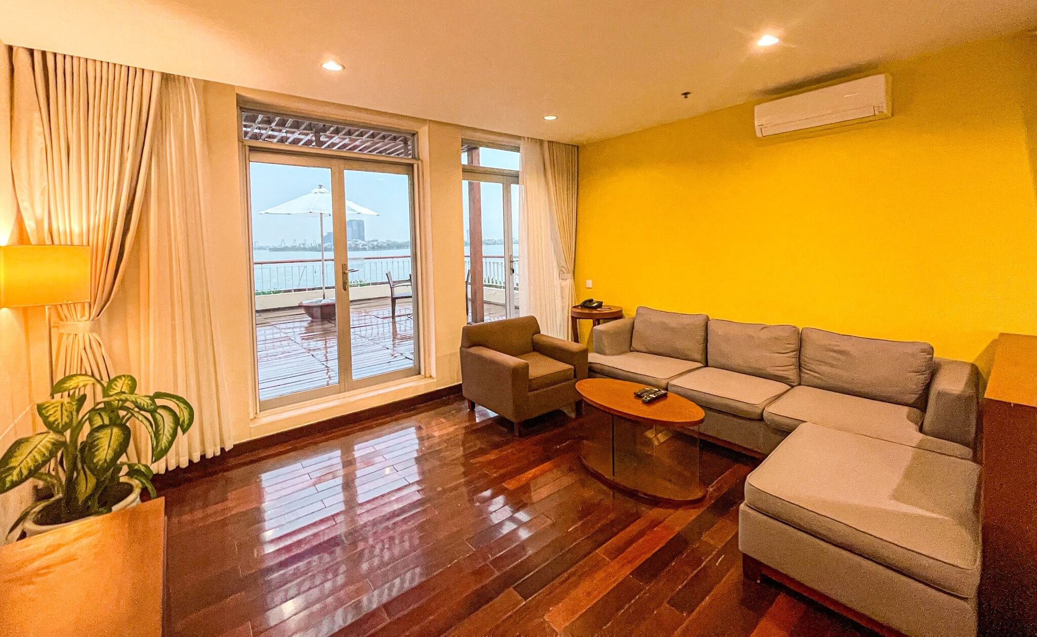 A living room with a sofa, armchair, and coffee table. There is a large window with curtains, a potted plant, and an air conditioning unit on the wall.